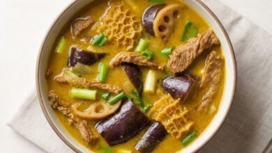 A bowl of Cambodian curry beef tripe soup with beef, eggplant, lotus shoots, and green onion in a coconut-curry broth.