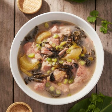 Text: Bowl of Cambodian Pork Hock with Pickle Mustard Green Soup on wooden table with soft natural light
