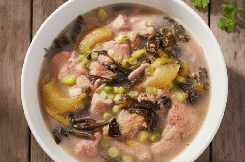 Text: Bowl of Cambodian Pork Hock with Pickle Mustard Green Soup on wooden table with soft natural light