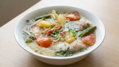 Overhead view of Cambodian sweet sour fish soup with fish chunks, pineapple, okra, tomatoes, bean sprouts, and fresh herbs in a serving bowl.