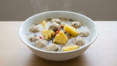 A bowl of Cambodian fish ball soup with pineapple chunks, fresh herbs, and fried garlic in a clear broth.