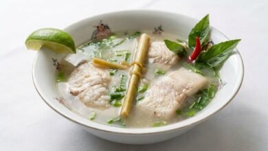 Overhead view of Khmer lemony fish soup with fish chunks, rice, green onion, herbs, and lime slices in a bowl.