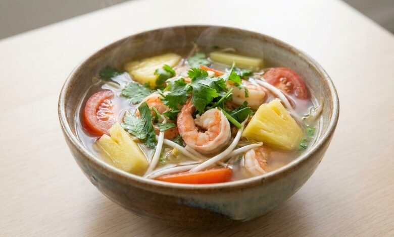 A bowl of Cambodian sweet sour and spicy shrimp soup with shrimp, pineapple, tomato, bean sprouts, and fresh herbs.