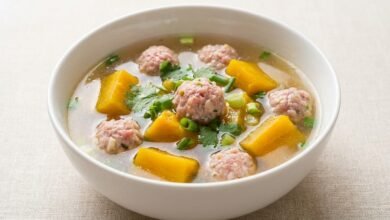 A bowl of Cambodian pumpkin and pork meatballs soup with visible meatballs, pumpkin cubes, and green onion in a clear broth.