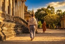 A traveler dressed modestly walks through a Cambodian temple grounds at golden hour with a monk in the background