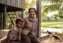 An elderly Cambodian woman sits with her young grandchild on a wooden platform beneath a rural stilt house.