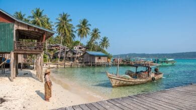 A wooden fishing boat returns to a small pier on a Cambodian island at dawn, with a woman in a krama waiting on the shore.