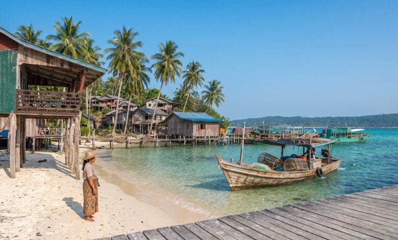 A wooden fishing boat returns to a small pier on a Cambodian island at dawn, with a woman in a krama waiting on the shore.