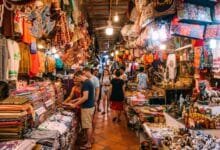 A traveler browsing colorful hand-woven silk scarves at a market stall in Cambodia with a smiling vendor