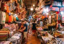 A traveler browsing colorful hand-woven silk scarves at a market stall in Cambodia with a smiling vendor