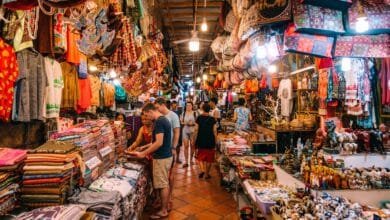 A traveler browsing colorful hand-woven silk scarves at a market stall in Cambodia with a smiling vendor