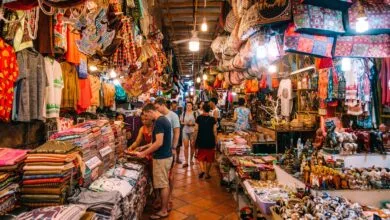 A traveler browsing colorful hand-woven silk scarves at a market stall in Cambodia with a smiling vendor