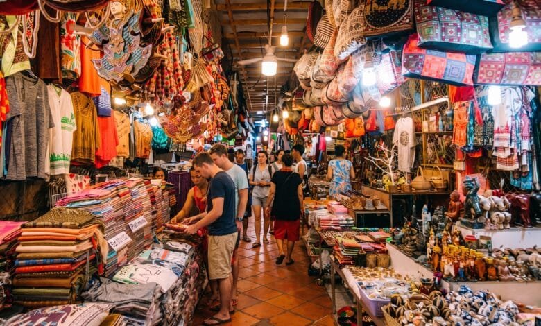 A traveler browsing colorful hand-woven silk scarves at a market stall in Cambodia with a smiling vendor
