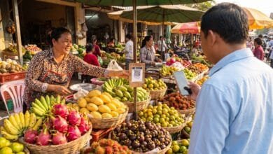 A vendor at a Cambodian market displays a QR code payment placard while a customer pays with a smartphone.