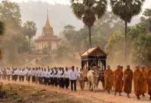 A line of mourners dressed in white walk behind a funeral cart toward a Cambodian pagoda.