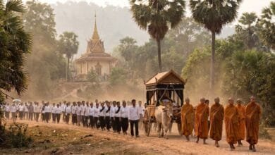 A line of mourners dressed in white walk behind a funeral cart toward a Cambodian pagoda.