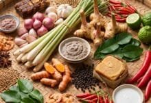 Fresh lemongrass, galangal, turmeric, kaffir lime leaves, prahok paste, Kampot pepper, and palm sugar arranged on a wooden table.