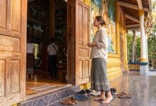 A traveler standing respectfully at the entrance of a Cambodian pagoda, shoes removed, looking toward the main hall