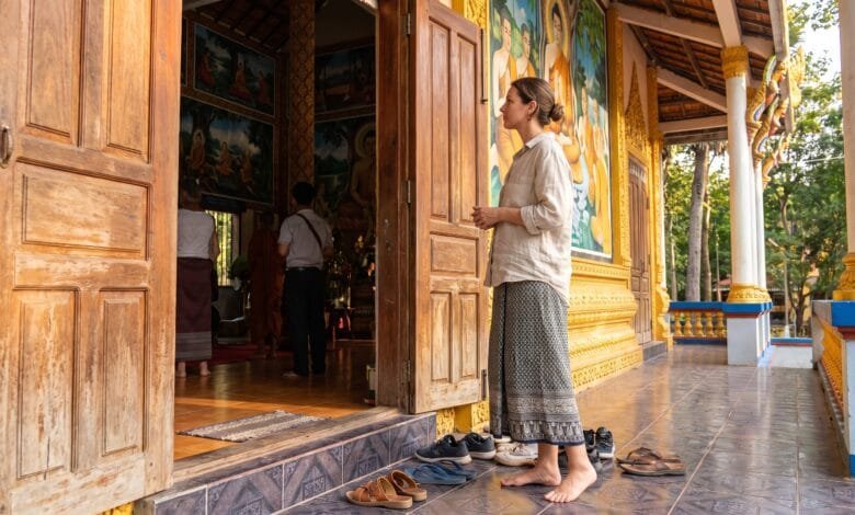 A traveler standing respectfully at the entrance of a Cambodian pagoda, shoes removed, looking toward the main hall