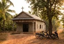 A small church building with a cross on the roof, surrounded by tropical vegetation in Cambodia