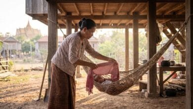 A Cambodian woman gently places a traditional krama scarf over a baby sleeping in a hammock outside her home.