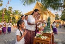 A Cambodian woman pours perfumed water over a Buddha statue at a pagoda during Khmer New Year, with a young girl watching respectfully beside her.