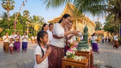 A Cambodian woman pours perfumed water over a Buddha statue at a pagoda during Khmer New Year, with a young girl watching respectfully beside her.