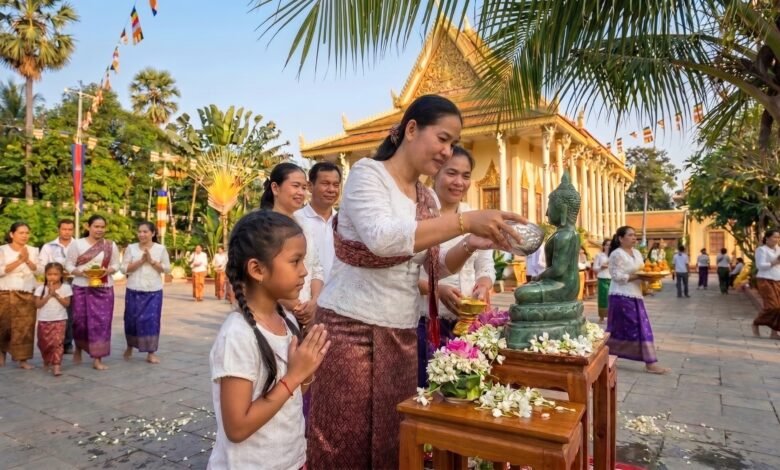 A Cambodian woman pours perfumed water over a Buddha statue at a pagoda during Khmer New Year, with a young girl watching respectfully beside her.