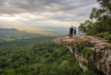 A man and woman stand on a rocky cliff overhang looking out over a vast green forest valley under a cloudy sky in Kirirom National Park, Cambodia.