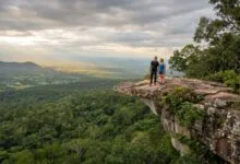 A man and woman stand on a rocky cliff overhang looking out over a vast green forest valley under a cloudy sky in Kirirom National Park, Cambodia.