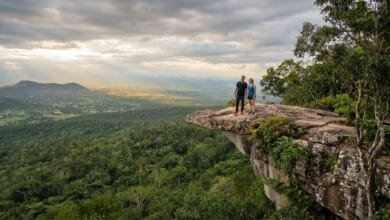 A man and woman stand on a rocky cliff overhang looking out over a vast green forest valley under a cloudy sky in Kirirom National Park, Cambodia.