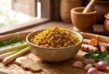 Golden-yellow Cambodian kroeung spice paste in a ceramic bowl surrounded by lemongrass, galangal, turmeric, and kaffir lime leaves.