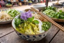 A bowl of Cambodian num banh chok with fresh rice noodles, green fish curry sauce, and raw vegetables at a street food stall