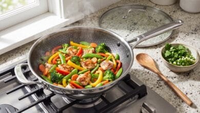OXO Hybrid 12-inch nonstick wok on a stovetop filled with a colorful vegetable stir-fry, with its glass lid resting on the counter nearby.
