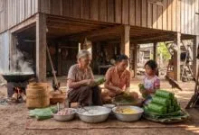 Elderly Cambodian grandmother wrapping banana leaves around Num Ansom sticky rice cake as her daughter and granddaughter watch in a traditional village kitchen