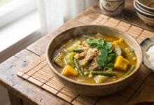 A bowl of Cambodian Samlor Korko soup with vegetables, served with jasmine rice on a wooden table.