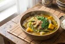 A bowl of Cambodian Samlor Korko soup with vegetables, served with jasmine rice on a wooden table.