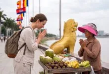 A foreign traveler presses their palms together in a sampeah gesture toward a smiling Cambodian vendor at a riverside stall near the Royal Palace in Phnom Penh.