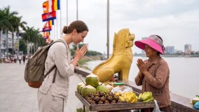 A foreign traveler presses their palms together in a sampeah gesture toward a smiling Cambodian vendor at a riverside stall near the Royal Palace in Phnom Penh.
